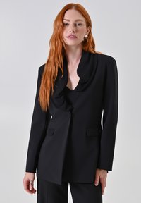 Blazer nero su misura con colletto drappeggiato, due tasche frontali e chiusura con un solo bottone. Texture liscia e design strutturato.