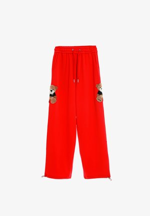 Pantalones de chándal rojos con un corte relajado, cintura con cordón y dos bolsillos decorados con apliques de oso de peluche. Textura suave con cremalleras laterales.