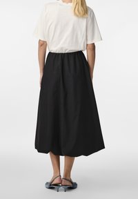 Frau trägt ein weißes kurzärmeliges Oberteil und einen taillenhohen, A-Linien schwarzen Midirock mit grauen Slingback-Kitten-Heels und steht mit dem Rücken zur Kamera.