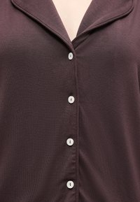 Chemise marron foncé à boutons, avec quatre boutons blancs visibles et une ouverture col en haut, portée par une personne.