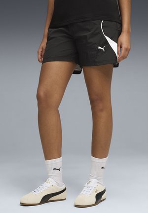 Person trägt schwarze Puma-Shorts, weiße Puma-Socken und weiße Puma-Sneaker und steht vor einem grauen Hintergrund.