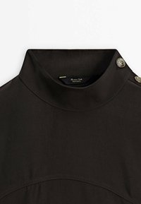 Blusa nera con colletto alto in tessuto liscio, caratterizzata da bottoni decorativi sulla spalla e dettagli di cuciture curve sottili.