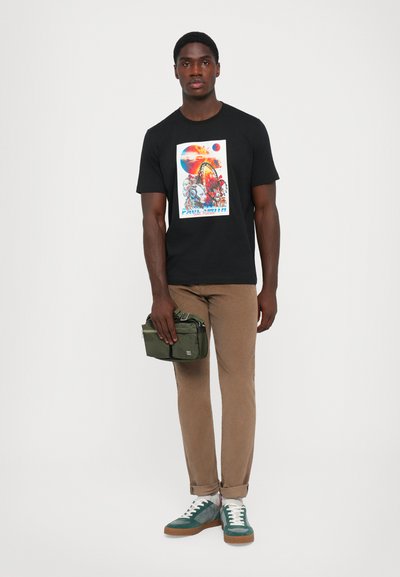 Sort bomulds t-shirt med et farverigt grafisk print, beige bukser, grøn taske og teal sneakers med hvide snørebånd.