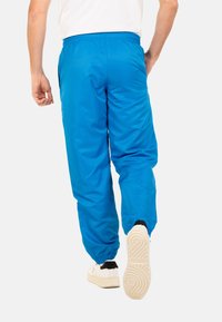 Blaue Sporthose aus leichtem Material mit elastischem Bund, elastischen Bündchen und glatter Textur. Getragen mit weißen Schuhen.