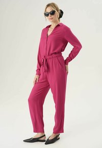 Tuta fucsia con maniche lunghe e vita stretta. Presenta una chiusura frontale con bottoni, tasche laterali e gambe affusolate. Indossato con ballerine nere.