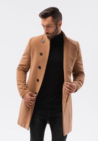 Cappotto in misto lana color tan con bottoni neri, colletto a risvolto, vestibilità slim e design monopetto, indossato sopra un dolcevita nero.