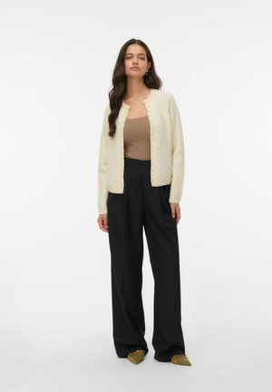 Crème gebreide cardigan met ronde halslijn, parelachtige knopen en een zachte textuur, gecombineerd met een beige top en brede zwarte broeken.