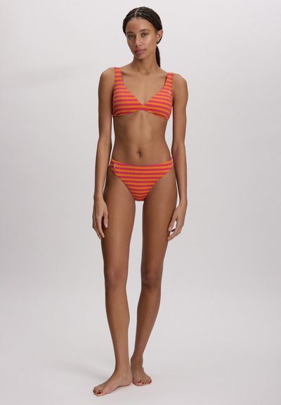 Γυναίκα που στέκεται ξυπόλητη φορώντας ένα ροζ και πορτοκαλί ριγέ μπικίνι μπροστά σε απλό, ανοιχτό γκρι φόντο.