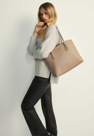 Beżowa skórzana torba typu tote z dwoma uchwytami u góry, zamknięciem na zamek błyskawiczny oraz minimalistycznym złotym logo. Nosi się ją z jasnoszarym prążkowanym swetrem i czarnymi spodniami.