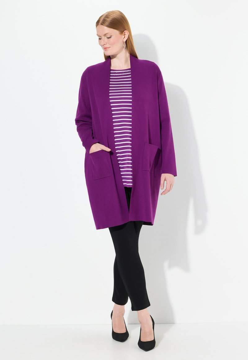 Lila Cardigan mit offenem Vorderteil, zwei vorderen Taschen, über einem gestreiften Top getragen, kombiniert mit schwarzen Leggings und hohen Schuhen. Glatte Textur.