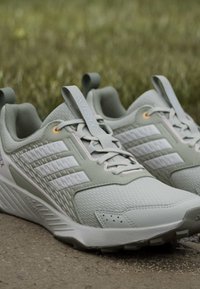 Ζευγάρι ανοιχτοπράσινα παπούτσια Adidas Terrex για τρέξιμο σε μονοπάτι με λευκές ρίγες σε χωμάτινο μονοπάτι, με φόντο θολό γρασίδι.