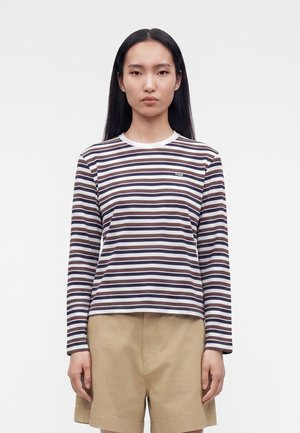 Ung kvinde med langt sort hår iført en langærmet navy og hvid stribet skjorte og beige shorts, stående mod en ensfarvet baggrund.