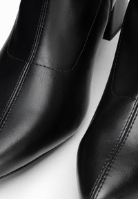 Gros plan de bottines en cuir noir à bouts pointus, coutures verticales et talons blocs, posées sur une surface blanche.