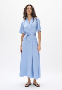 Hellblaues Wickelkleid aus weichem Stoff mit kurzen Ärmeln, einem Taillengürtel und einem langen, fließenden Rock. Kombiniert mit einfachen schwarzen Sandalen.