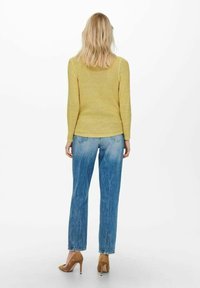 Femme aux cheveux blonds portant un pull en maille jaune, un jean bleu délavé et des chaussures à talons marron, debout de dos sur un fond blanc.