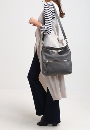 Žena ve pruhovaném topu a béžové vestě nese velkou černou koženou kabelku přes rameno s přední kapsou na zip.