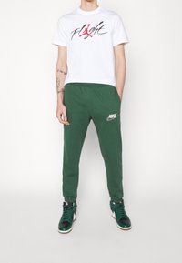Bílý tričko s černou a červenou grafikou "Flight", zelené teplákové kalhoty s bílým logem Nike a černo-zelené vysoké tenisky.