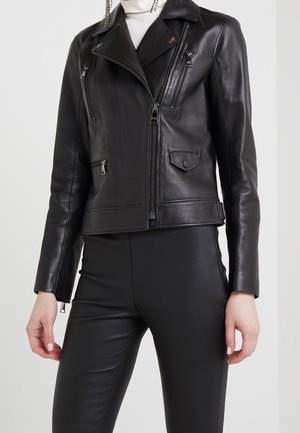 Femme portant une veste de biker en cuir noir avec des poches zippées et un pantalon noir ajusté, col roulé blanc visible au niveau du cou.