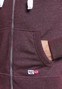 Sudadera con cremallera burdeos, con cordones blancos, bolsillos frontales y una pequeña etiqueta de logo roja y blanca en la parte inferior izquierda. Tejido texturizado.
