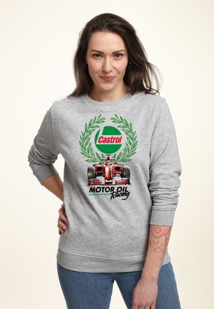 Femme portant un sweat-shirt gris avec une voiture de course rouge, une couronne de laurier verte et le texte "Castrol Motor Oil Racing", debout avec la main sur la hanche.