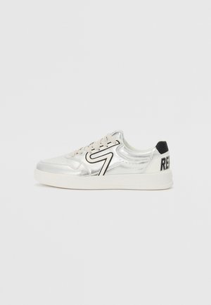 Baskets basses argentées avec semelle blanche, lacets beige, patch noir au talon arrière et motif zigzag noir sur le côté.