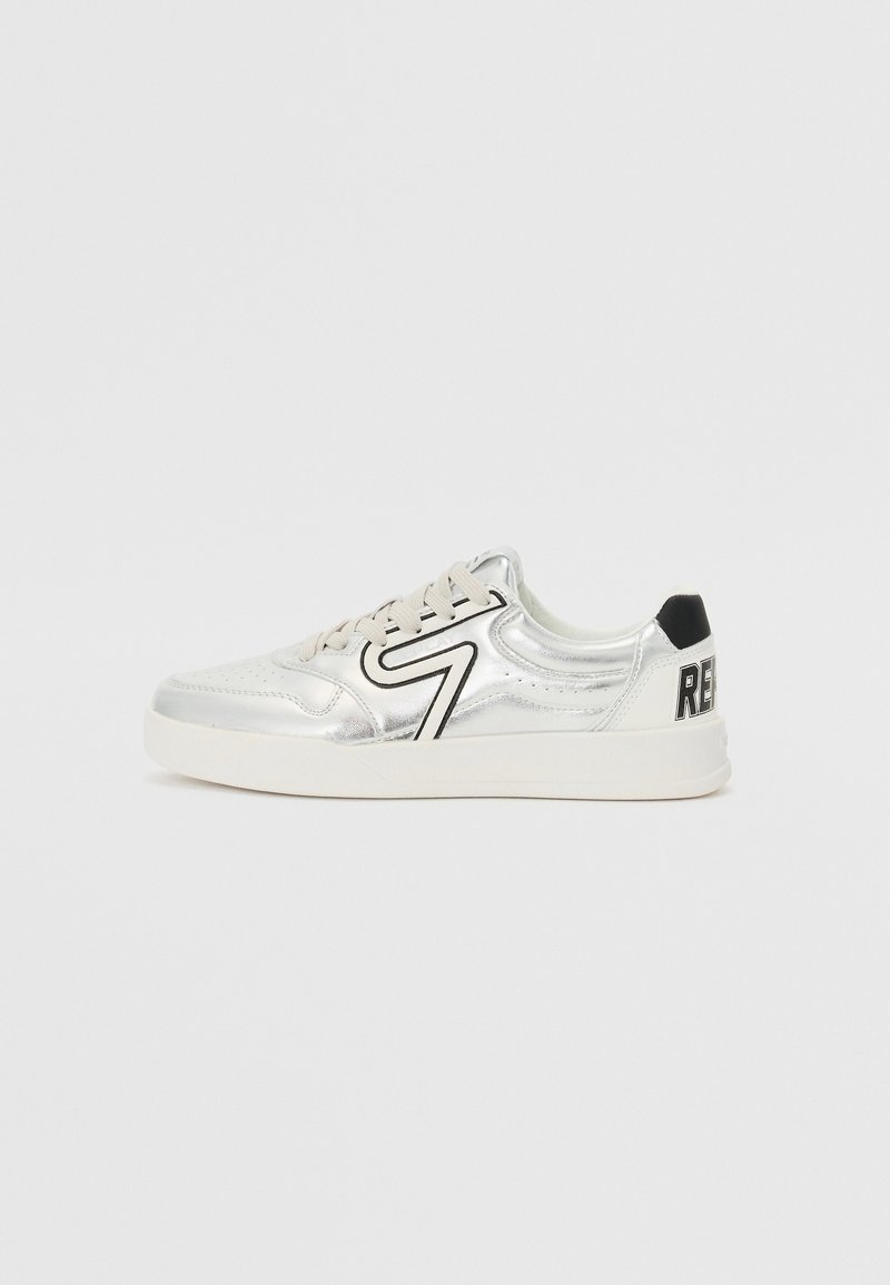 Baskets basses argentées avec semelle blanche, lacets beige, patch noir au talon arrière et motif zigzag noir sur le côté.