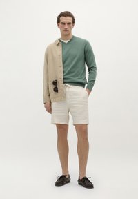 Licht beige knoopjack met groene V-halstrui, crèmekleurige shorts en donkerbruine schoenen. Model staat tegen een witte achtergrond.