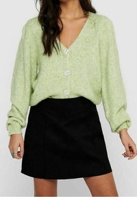 Hellgrüne Strickjacke mit V-Ausschnitt, drei großen Knöpfen und gepufften Ärmeln, kombiniert mit einem schwarzen Minirock. Weiche Textur und entspannte Passform.