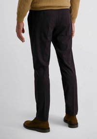 Mann in dunklen, schmal geschnittenen Hosen, braunen Wildleder-Stiefeletten und einem camel-farbenen, gerippten Pullover, der vor einem schlichten, hellen Hintergrund steht.