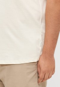 Helles cremefarbenes Baumwoll-T-Shirt mit geradem Schnitt, kurzen Ärmeln und einem seitlichen Etikett. Der Stoff fühlt sich weich und geschmeidig an und passt gut zu beigen Hosen.