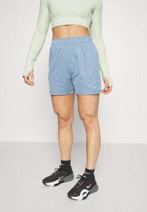 Personne portant un crop top à manches longues vert clair, un short de sport bleu clair, des baskets Nike noires et des chaussettes noires, debout sur un sol gris.