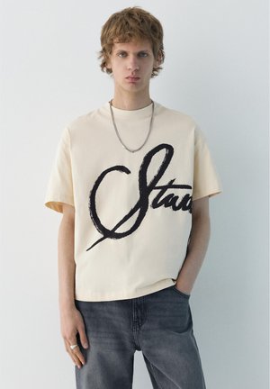 Mladý muž s krátkymi kučeravými vlasmi, oblečený v béžovom oversized tričku s veľkým čiernym nápisom a tmavosivých džínsach, stojaci pri bielej stene.