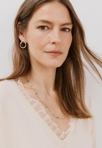 Lichtbeige trui met een V-hals en doorzichtige, gerimpelde afwerking. Gouden oorringen en een delicate halsketting van meerkleurige kralen voegen contrast toe.