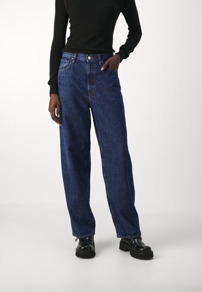 Jean en denim bleu taille haute avec une coupe ample. Présente une fermeture à bouton, des passants de ceinture et des poches avant. Porté avec des chaussures à plateforme noires.