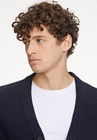 Mörkblå cardigan med ribbad krage, enstaka knappstängning, bärs över en vit t-shirt. Har en mjuk textur och avslappnad passform.