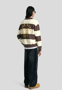 Hosszú, sötét hajú személy csíkos barna és krémszínű pulcsiban, fekete, bő nadrágban, barna csizmában és napszemüvegben áll oldalt.