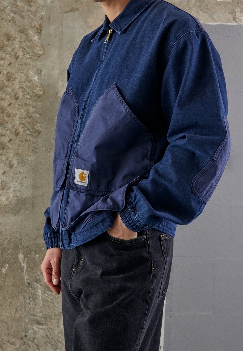 Veste en denim bleu marine avec poches contrastantes bleu marine, fermeture éclair et poignets élastiques. Comprend une étiquette avec le logo Carhartt sur la poche avant.