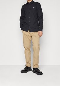 Camisa negra de manga larga con cuello abotonado, con un pequeño logo verde. Combinada con pantalones beige claros y zapatos negros.