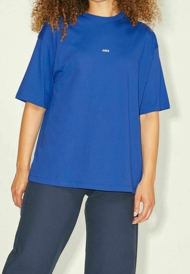 Modré oversized tričko vyrobené z bavlny s kulatým výstřihem a krátkými rukávy, s bílým logem na hrudi, doplněné tmavými kalhotami.