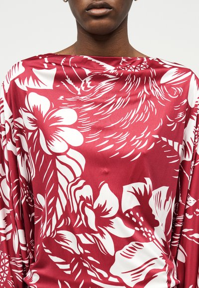 Γυναίκα που φοράει κόκκινο σατέν τοπ με μεγάλα λευκά λουλουδένια και αφηρημένα σχέδια, με λαιμόκοψη καραβόπανο και μακριά μανίκια.