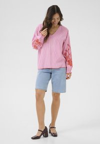 Růžový pruhovaný oversized top s dlouhými rukávy, zdobený červenou květinovou výšivkou, spárovaný se světle modrými džíny šortkami a tmavými botami na blokovém podpatku.
