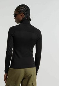 Maglietta nera a maniche lunghe con colletto alto, realizzata in tessuto a maglia di struttura, caratterizzata da un dettaglio sul retro. Indossata con pantaloni cargo verde oliva.