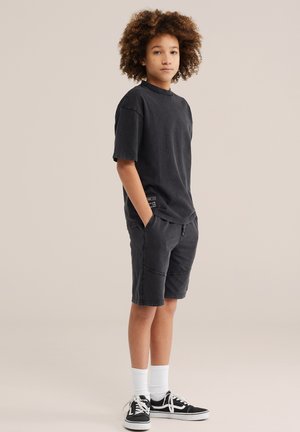 Nooruk lokkis juustega seisab käed taskutes, kannab musta ülemõõdulist t-särki, musti lühikesi pükse, valgeid sokke ja musti vabaajajalatsid.