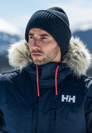 Marine winterjas met bont gevoerde capuchon, rode trekkoorden en logo op de borst. Geribde marinebeanie. Sneeuwachtige bergen op de achtergrond.