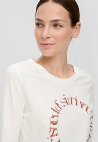 Koszula z długim rękawem w kolorze kremowym, z okrągłym dekoltem, z okrągłym brązowym wzorem tekstowym na przodzie. Miękka, lekka tkanina.