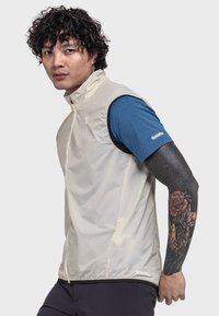 Beige, leichte Weste mit hohem Kragen, verfügt über eine strapazierfähige, glänzende Oberfläche. Wird über einem blauen Kurzarmhemd getragen. Enthält markenspezifische Akzente.