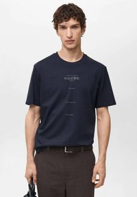 Navy blue cotton t-shirt with short sleeves, round neck, featuring text “GALERIE 1974” and other phrases printed in lighter ink on the front.
