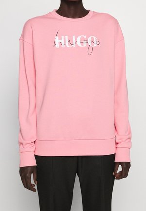 Person wearing a pink sweatshirt with the word "HUGO" in white and black script across the chest, paired with black pants.