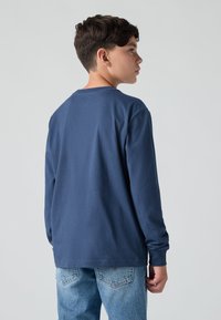 Sötétkék, hosszú ujjú póló kerek nyakkal és texturált anyagból, világoskék farmernadrággal párosítva. Hátulról nézve, laza fazonban.