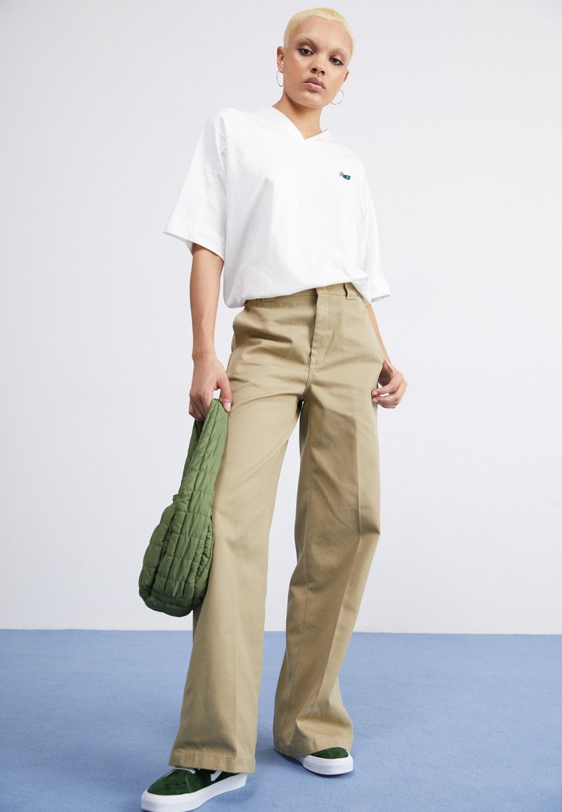 Weißes kurzärmeliges V-Ausschnitt-T-Shirt, beigefarbene weit geschnittene Hose, grüne steppierte Tasche und grüne Sneaker auf blauem Hintergrund.